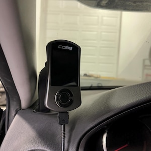 May include: A black and gray COBB tuning device mounted on a car's dashboard. The device has a screen and a central control button. A black cable is connected to the bottom of the device.
