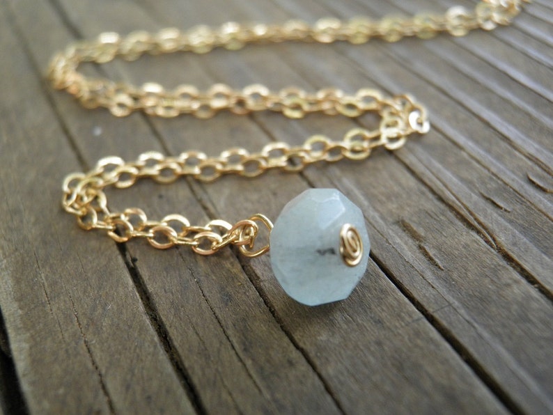 May include: A gold chain necklace with a light blue gemstone pendant. The pendant is faceted and has a gold wire wrapped around it.