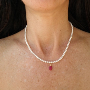 Può includere: Una collana di perle bianche con un ciondolo di pietra rossa.