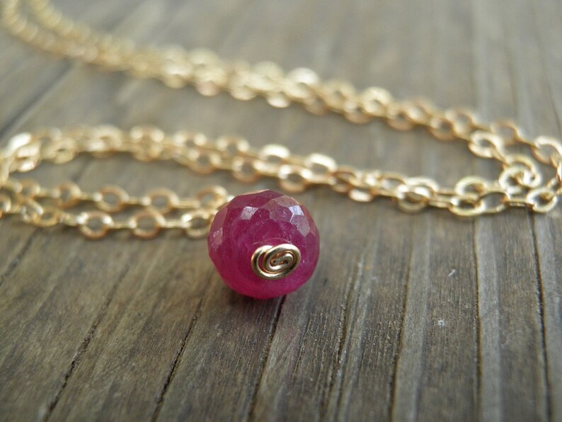 May include: A delicate gold chain necklace with a single, faceted, deep pink gemstone pendant.