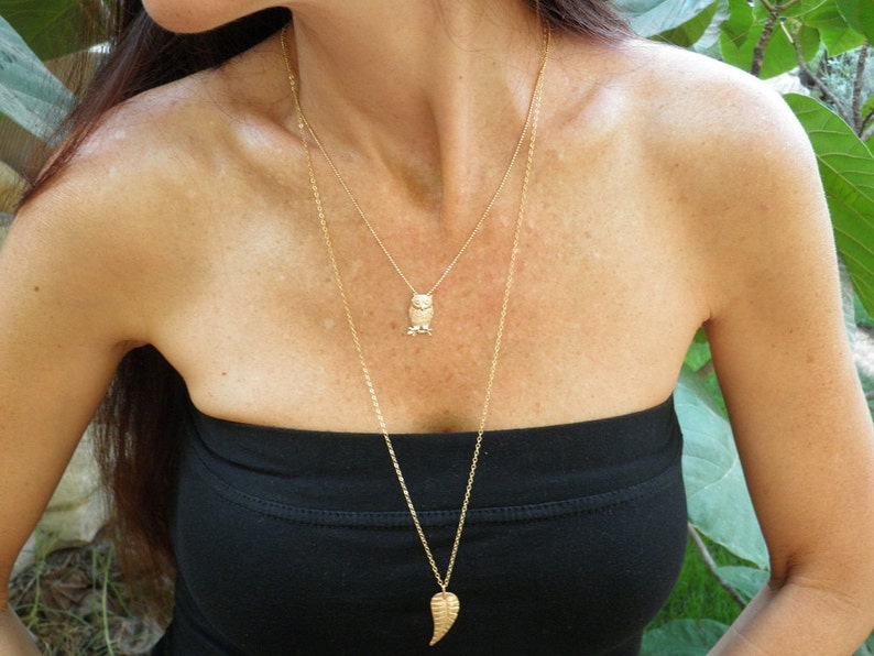 May include: A gold chain necklace with an owl pendant and a leaf pendant. The necklace is layered and worn by a person with long brown hair.
