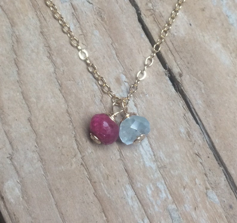 May include: A gold chain necklace with two gemstone pendants. The pendant on the left is a deep pink, and the pendant on the right is a light blue.