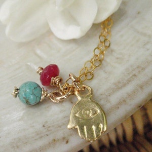 May include: Gold chain necklace with a turquoise and red stone charm and a gold hamsa hand charm with an evil eye detail.