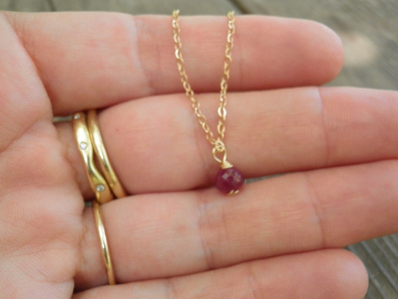May include: A delicate gold chain necklace with a small, round, dark red gemstone pendant.