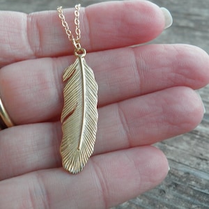 Puede incluir: Un collar de oro con un colgante de pluma. La pluma es detallada y tiene una superficie texturizada.