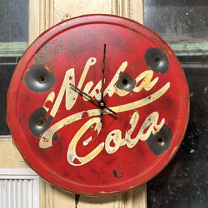 May include: A round, red clock with the words "Nuka Cola" in white and yellow script. The clock face has a distressed, vintage look with several bullet holes. The clock hands are black.