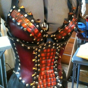 May include: A handcrafted leather corset with a striking design. The corset features a combination of black and burgundy leather, accented with a textured, orange-red pattern and silver studs. The design includes straps and a decorative buckle.