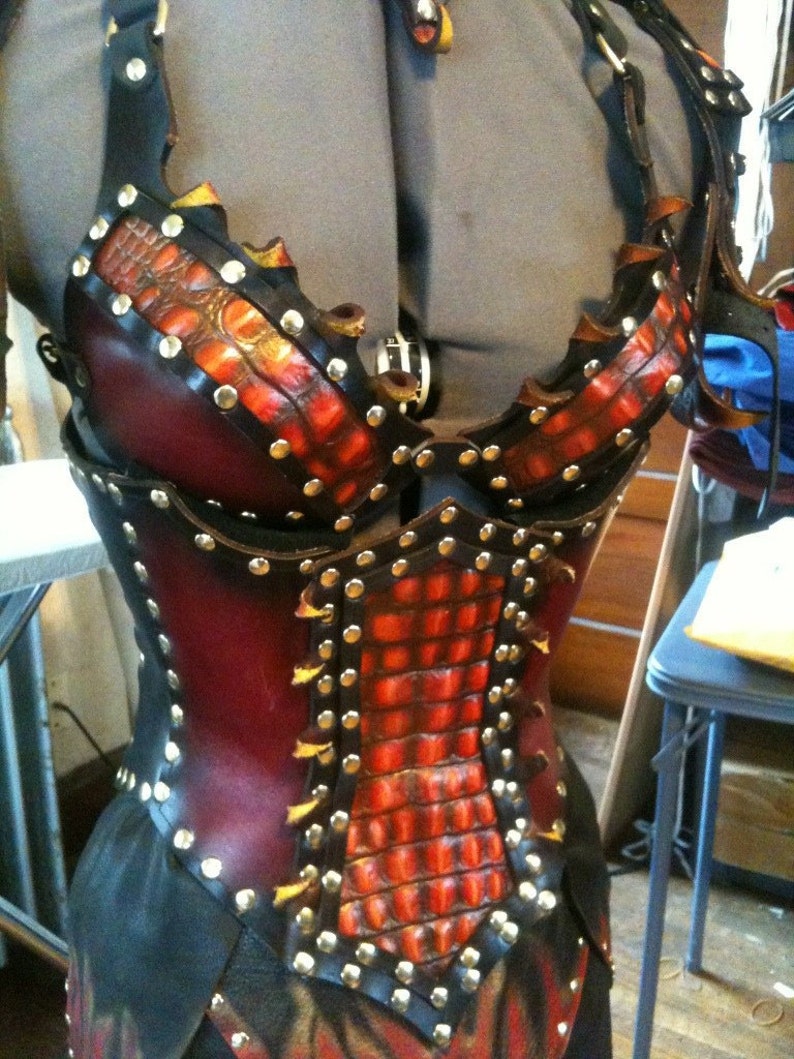May include: A handcrafted leather corset with a striking design. The corset features a combination of black and burgundy leather, accented with a textured, orange-red pattern and silver studs. The design includes straps and a decorative buckle.
