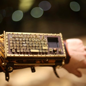 May include: A steampunk-style wrist-mounted keyboard with a small screen and brass accents. The keyboard features small, square keys and is attached to a brown leather wristband with decorative etching. The overall design is intricate and detailed.