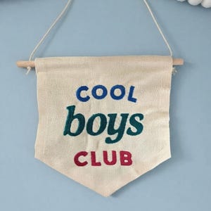 May include: A cream-coloured fabric wall banner with a pointed bottom edge, suspended from a wooden dowel by a white string. The banner features embroidered text in three colours: COOL in blue, boys in dark green, and CLUB in red, centred on the fabric.