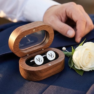 以下が含まれることがあります： 白い背景にイニシャル「MG」が入った2つのシルバーカフスボタンが入った、透明な蓋付きの木製の楕円形の箱。白いバラのブートニアと人の手は背景にあります。