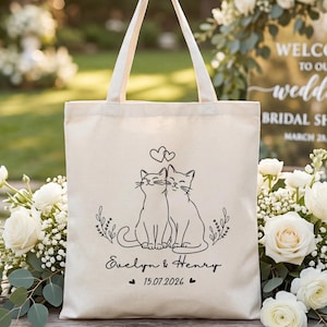 May include: A cream-colored tote bag with black line art of two cats under two hearts. The bag has the names "Evelyn & Henry" and the date "15.07.2026" printed on it. The bag is surrounded by white flowers.