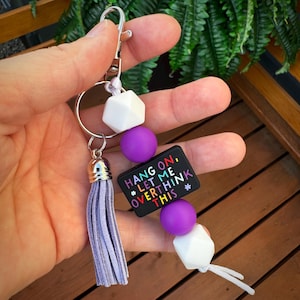 May include: A keychain with a silver clasp, a purple tassel, and a beaded design. The beads are white geometric shapes and purple spheres. A black rectangular charm reads "HANG ON, *LET ME OVERTHINK THIS*" in colorful text.