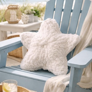 Puede incluir: Un gran coj&iacute;n blanco con forma de estrella de mar descansa sobre una silla Adirondack azul claro. La silla est&aacute; en una terraza con vista a una playa y al oc&eacute;ano. Una manta blanca est&aacute; sobre la silla. Una bandeja con un vaso de agua y rodajas de lim&oacute;n est&aacute; cerca.