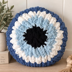Puede incluir: Un cojín redondo de punto grueso con un diseño de ojo concéntrico. El centro es un círculo negro sólido, rodeado por anillos de color azul claro, blanco y un borde exterior grueso de hilo azul oscuro, creando una apariencia texturizada y suave.