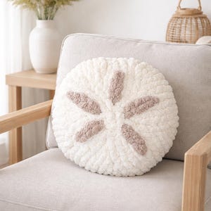 May include: A round, cream-colored decorative pillow with a sand dollar design. The pillow is made of a soft, textured material and features six light brown petal-shaped accents. It rests on a light gray armchair.