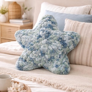 Puede incluir: Un gran coj&iacute;n en forma de estrella en tonos azules y verdes, colocado sobre una cama. El coj&iacute;n tiene un aspecto texturizado y afelpado, lo que sugiere un material suave y c&oacute;modo. El entorno del dormitorio incluye otros cojines y una mesita de noche.
