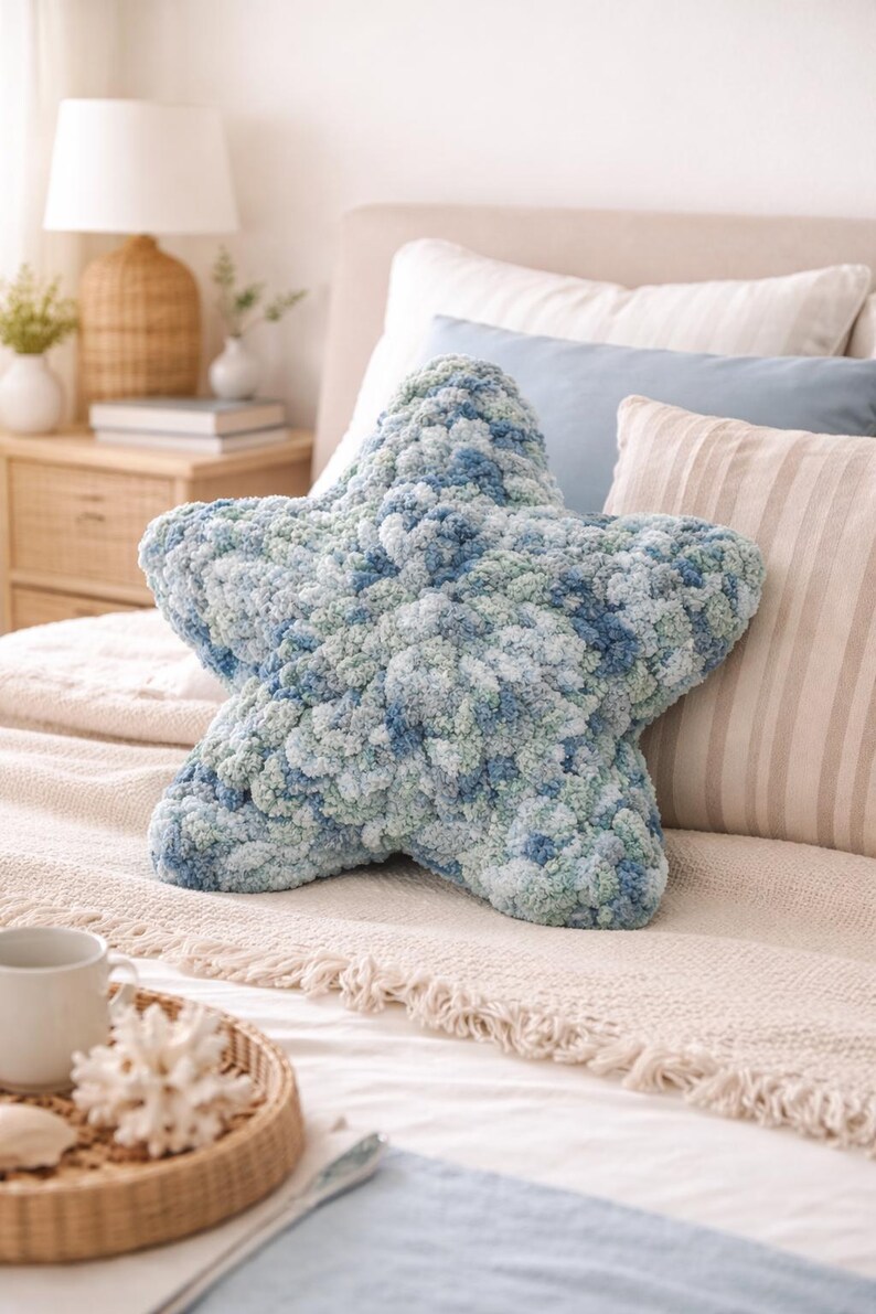 Puede incluir: Un gran coj&iacute;n en forma de estrella en tonos azules y verdes, colocado sobre una cama. El coj&iacute;n tiene un aspecto texturizado y afelpado, lo que sugiere un material suave y c&oacute;modo. El entorno del dormitorio incluye otros cojines y una mesita de noche.