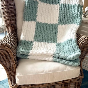 May include: A chunky knit blanket in a checkerboard pattern of white and seafoam green rests on a wicker chair with a white cushion. The blanket's thick yarn creates a cozy, textured appearance, perfect for home decor.