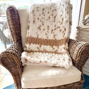 Puede incluir: Una manta de punto grueso en blanco y beige, colocada sobre una silla de mimbre con un cojín blanco. La manta presenta un diseño a rayas con una banda beige sólida en el medio. La silla es de mimbre marrón tejido.