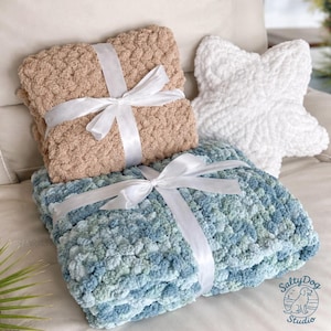 Puede incluir: Un conjunto de tres artículos de felpa: una manta beige, una manta azul y verde, y un cojín blanco en forma de estrella de mar. Cada manta está atada con una cinta de raso blanca. Las mantas tienen un aspecto texturizado y nudoso.