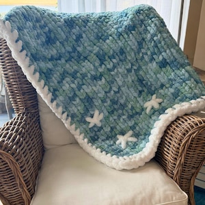 Puede incluir: Una manta tejida a mano, en tonos azules y verdes, adornada con aplicaciones de estrellas de mar blancas. La manta tiene un borde blanco y esponjoso y está colocada sobre una silla de mimbre con un cojín blanco. El logotipo de Salty Dog Studio es visible en la esquina superior izquierda.