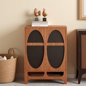May include: A wooden cabinet with two oval-shaped, dark-colored panels on the doors. The cabinet has a light brown finish and is adorned with two bird sculptures on top. A woven basket is on the left.