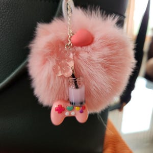 May include: A pink fluffy pom-pom keyring with a silver chain. The keyring features a pink heart, a clear gummy bear, a small drink charm, and a pink game controller charm with colourful buttons. The background is a dark green handbag.