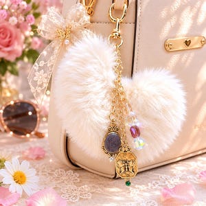 May include: A cream-colored handbag with gold hardware and a fluffy, heart-shaped keychain. The keychain features gold charms, including a cameo and a bow, and is adorned with colorful beads. A lace bow with a butterfly accent decorates the bag.