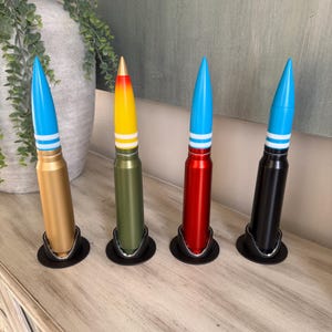 May include: Four decorative artillery shell replicas in gold, olive green, red, and black, each with a blue tip and white stripes. They are displayed in black holders on a wooden surface.
