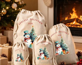 May include: Three beige drawstring gift bags made of a hessian-style fabric. Each bag features a printed watercolour illustration of a snowman wearing a blue hat and scarf next to a Christmas tree, with the text Special Delivery For printed above.