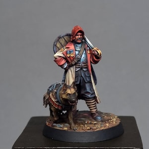 May include: Miniature figurine of a man in a red coat and hat, holding a sword and shield, with a dog wearing a harness. The figure is standing on a small, round base. The dog is a German Shepherd.