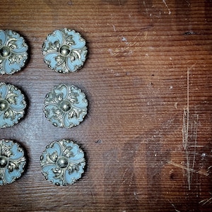 May include: Six antique, round cabinet knobs with a light blue and gold floral design. The knobs are arranged on a dark brown, textured wooden surface. Each knob features a central, raised gold detail.