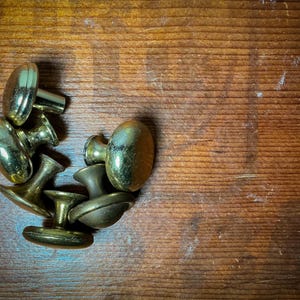 Puede incluir: Un conjunto de pomos de puerta antiguos de latón con forma ovalada. Los pomos están dispuestos sobre una superficie de madera, mostrando su acabado dorado envejecido. Los pomos tienen una estética clásica y vintage.