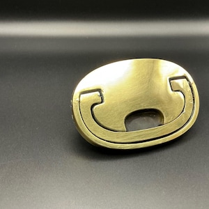May include: Oval-shaped, antique brass-colored belt buckle. The buckle features a unique design with curved edges and a central opening. The metal has a slightly worn appearance, suggesting an aged or vintage aesthetic.