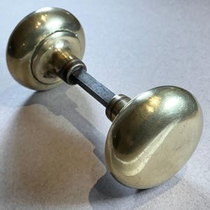 May include: A vintage brass door knob set. The set features two oval-shaped knobs connected by a dark metal rod. The knobs have a polished, reflective surface, and the overall design suggests a classic or antique style.