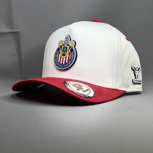 Peut inclure: Casquette de baseball blanche et rouge avec le logo du Club Deportivo Guadalajara. La casquette a une visière et un bouton supérieur rouges, avec l'emblème de l'équipe à l'avant. Le mot "Chivas" est également présent sur le côté.