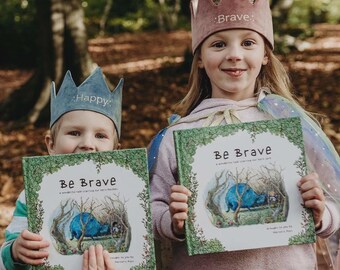 Puede incluir: Dos niños con coronas bordadas con las palabras "Brave" y "Happy" sostienen libros titulados "Be Brave: Un cuento maravilloso que celebra nuestra fuerza interior".