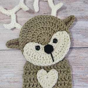 May include: A handmade crochet deer with a brown body, cream-colored face, and white antlers. The deer has black button eyes, a black nose, and a cream-colored heart on its chest. The deer's feet are brown.