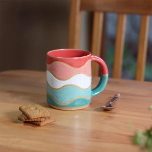 Op de afbeelding: Een keramische mok met een golvend ontwerp in koraal, roze, wit en blauwgroen. De mok heeft een blauwgroene handgreep en een bijpassende blauwgroene basis. Een kleine lepel en een stapel koekjes liggen op de houten tafel.