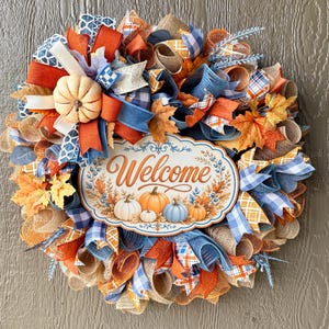 Può includere: Una ghirlanda decorativa con un cartello ovale centrale con il testo Welcome, circondata da nastri in rete arancioni, blu e marrone chiaro, strisce di tessuto a quadri, foglie d'acero artificiali e un piccolo accento a zucca bianca.
