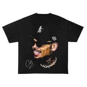 May include: Black t-shirt featuring a portrait of a smiling person wearing sunglasses and a baseball cap. The person has tattoos and a gold chain. The shirt has a signature on the lower left.
