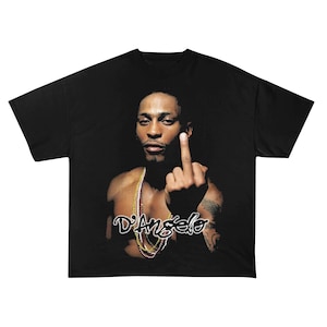 Può includere: T-shirt nero con un ritratto di D'Angelo con una collana d'oro, che mostra il dito medio. Il testo "D'Angelo" è scritto sotto. La maglietta ha una vestibilità ampia ed è in tessuto scuro.