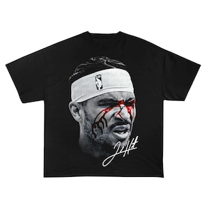 Puede incluir: Camiseta negra con un retrato en escala de grises de una persona con rayas rojas en la cara. La persona lleva una cinta blanca con el logo de la NBA. La firma "JH" es en blanco.