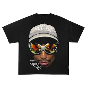 May include: Black t-shirt with a portrait wearing sunglasses and a cap. The cap says "Human Made" in white. The sunglasses reflect an orange and yellow scene. The shirt has a white signature.
