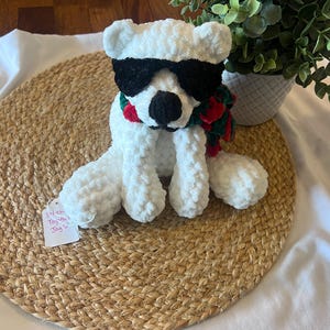 May include: A white plush polar bear toy wearing black sunglasses and a red, green, and white scarf. The bear is sitting on a woven mat. A small tag is attached to the bear. A potted plant is in the background.