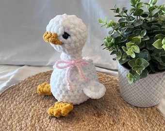 Puede incluir: Un ganso de crochet blanco hecho a mano con pico y patas amarillas, y un lazo rosa. El peluche está sobre una estera tejida, con una planta en maceta en el fondo. El ganso es un objeto decorativo encantador.