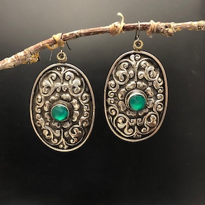 May include: A pair of silver earrings with an oval shape and intricate floral designs. Each earring features a green gemstone in the center.