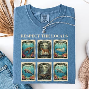 May include: Blue Comfort Colors t-shirt with the text "Respect the Locals." The shirt features six vintage-style postage stamps with illustrations of sea creatures and animals, including a sea turtle and panther.