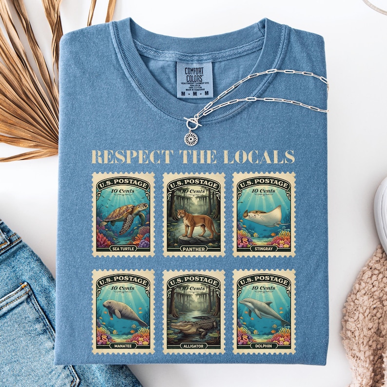 May include: Blue Comfort Colors t-shirt with the text "Respect the Locals." The shirt features six vintage-style postage stamps with illustrations of sea creatures and animals, including a sea turtle and panther.